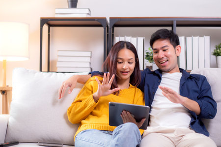 Happy asian couple video call on tablet sitting on sofa in living room at home, smiling and waving during online communication with relax, couple watching tablet, lifestyle and interaction.の写真素材
