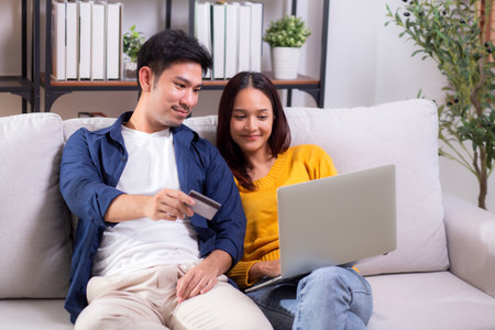 Happy asian couple using laptop and credit card for online shopping together in living room at home, man and woman smiling and relax with enjoy a digital lifestyle, convenient payment.の写真素材