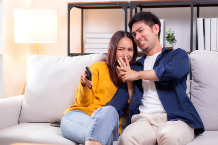 Young asian couple watching a scary or thrilling TV show in living room at home with emotion shock and scared, emotion reaction while watching horror television with fear, lifestyle and bonding.の写真素材