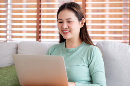 Portrait young asian business woman sitting on sofa using laptop computer in living room at home, businesswoman sitting on couch working on notebook with comfortable, business and freelance.の写真素材