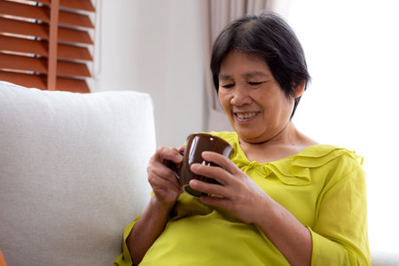 Happiness senior woman sitting on sofa and holding a coffee cup drinking looking out a window in living room at home, elderly drinking beverage for relax, lifestyle and cozy, domestic life.の写真素材