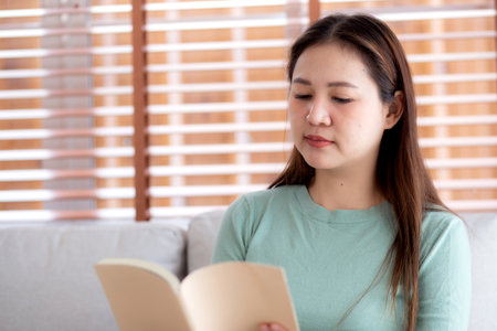 Young asian woman reading a book with expression sitting on sofa in living room at home, woman relax and leisure sitting on couch reading literature, activity and hobby, lifestyle and relaxation.の写真素材