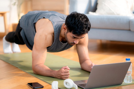 Young asian man doing plank exercise in living room at home while watching online workout tutorial on laptop with endurance and motivation, man training plank on yoga mat, digital fitness lifestyle.の写真素材