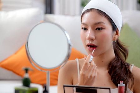 Beauty happy young asian woman applying makeup with lipstick looking mirror in living room at home,...