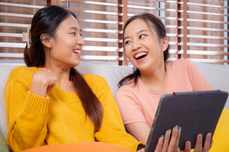 Two young asian woman sitting on sofa using digital tablet for video call in living room at home,...