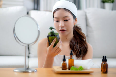 Beautiful young asian woman cheerful holding skincare or cream herb sitting on sofa at home, beauty woman hold skin care or lotion for rejuvenation on facial, cosmetic and makeup for self care.の写真素材