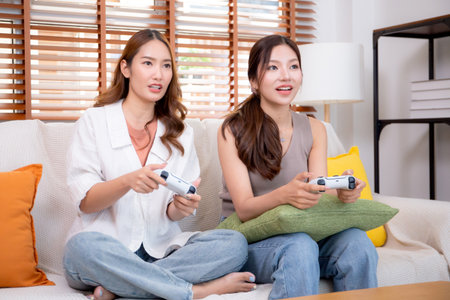 Two asian woman playing video games together with controllers in a cozy living room at home, fun,...