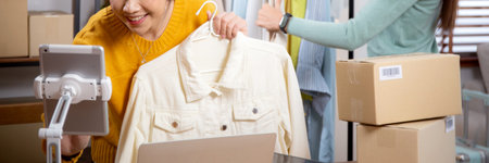 Two young asian woman live online clothing store while showing clothing with tablet and laptop on social at home, entrepreneur streaming for selling online clothing and presenting, online seller.の写真素材