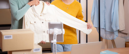 Two young asian woman live online clothing store while showing clothing with tablet on social at home, entrepreneur streaming for selling online clothing and presenting, online seller.の写真素材