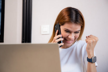 Young caucasian businesswoman talking with smartphone and working on laptop with glad in office,...