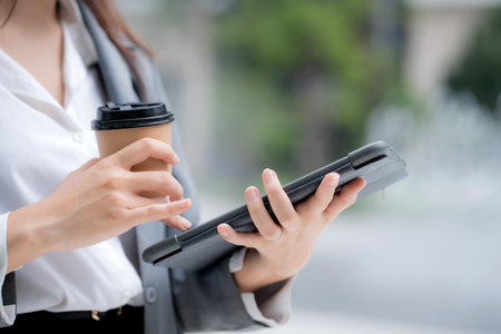 Close up hand of businesswoman standing holding coffee while using tablet outdoors city, digital nomad, business woman or manager touching tablet for working, modern technology, digital device.の写真素材