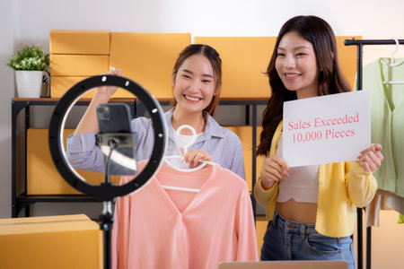 Two asian woman live streaming online clothing store while showing clothing with smartphone on social and success, entrepreneur streaming for selling online clothing and presenting, online seller.の写真素材