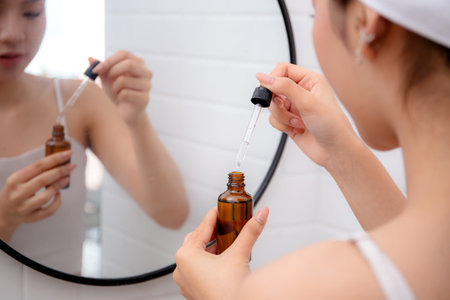 Beautiful young asian woman applying serum for rejuvenation and looking mirror in bathroom, beauty woman applying collagen for treatment skin, self-care, skincare and makeup, cosmetic and makeup.の写真素材