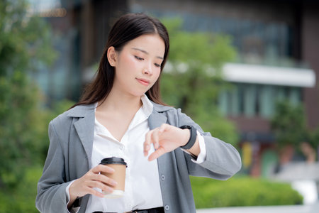 Young asian businesswoman checking time on smartwatch while waiting on busy city street, professional business woman with confident smile managing schedule and appointment.の写真素材