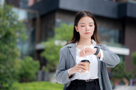 Young asian businesswoman checking time on smartwatch while waiting on busy city street with stress and worried, professional business woman managing schedule and appointment in business district.の写真素材