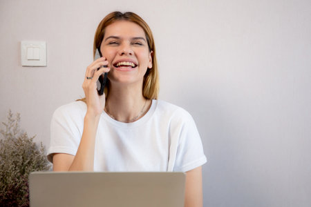 Young caucasian businesswoman talking with smartphone and working on laptop computer in home office, business woman working on laptop and speaking on smart phone, business and communication.の写真素材