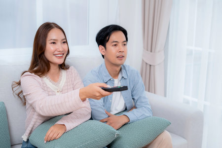 Happy asian couple watching television together sitting on sofa in living room at home, using remote...