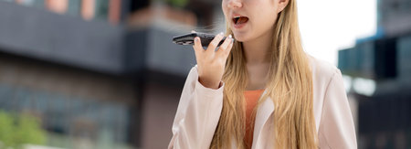 Young caucasian businesswoman using voice message on smartphone with chatbot or AI outdoors in city,...