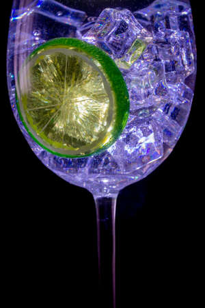Stemmed glass with clear liquid, lime slice, and ice cubes against black backdrop.の写真素材