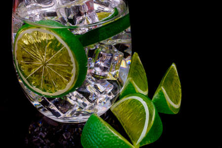 Glass with clear liquid, ice, and lime slice on black reflective acrylic.の写真素材