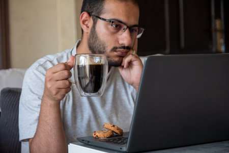 bearded male drinks coffee while working on his laptop from his home and eating cookiesの写真素材