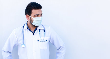 Young male wearing mask and doctor coat while holding stethoscope on his neck in confident wayの写真素材