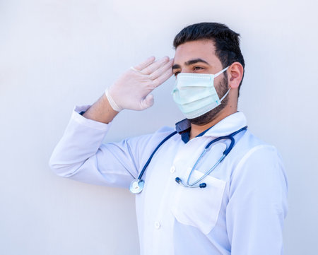 Doctor doing military salute on isolated white background holding his stethoscope wearing mask during healthcare dayの写真素材