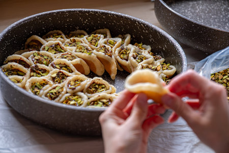 Hands holding Qatayef filled with chocolate and topped with pistachios on a wooden table, with a plate to be prepared in the oven later as Ramadan sweets.の写真素材
