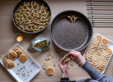 Hands holding Qatayef filled with chocolate and topped with pistachios on a wooden table, with a plate to be prepared in the oven later as Ramadan sweets.の写真素材