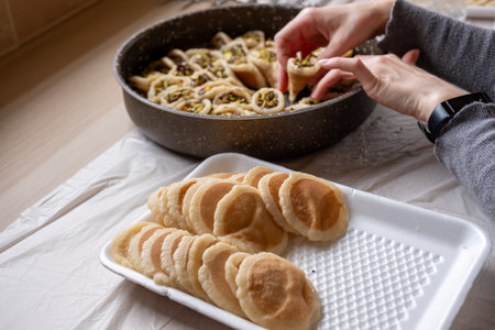 Hands holding Qatayef filled with chocolate and topped with pistachios on a wooden table, with a plate to be prepared in the oven later as Ramadan sweets.の写真素材