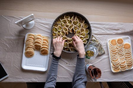 Hands holding Qatayef filled with chocolate and topped with pistachios on a wooden table, with a plate to be prepared in the oven later as Ramadan sweets.の写真素材