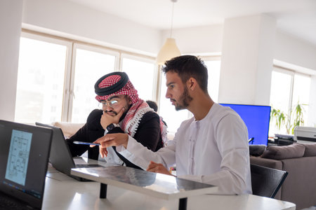 Business Meeting Between Colleagues in Modern Office, One in Traditional Arab Attire, Using Laptop...