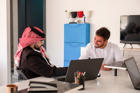 Two men share a friendly business meeting in a bright, modern office. One wears traditional Middle...