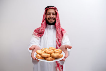In a gesture of hospitality and generosity, an Arabic man proudly presents a plate of Maamoul, symbolizing the warmth and abundance of Eid celebrations. Clad in the traditional Kandura and Keffiyehの写真素材