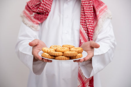 In a gesture of hospitality and generosity, an Arabic man proudly presents a plate of Maamoul, symbolizing the warmth and abundance of Eid celebrations. Clad in the traditional Kandura and Keffiyehの写真素材
