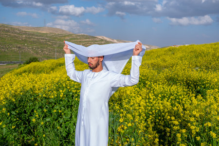 A man in a white dishdasha and kandura stands in a field of yellow flowers, lifting the kandora with eyes closed, enjoying the spring sunshine, surrounded by green hills under a partly cloudy skyの写真素材