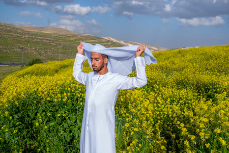A man in a white dishdasha and kandura stands in a field of yellow flowers, lifting the kandora with eyes closed, enjoying the spring sunshine, surrounded by green hills under a partly cloudy skyの写真素材