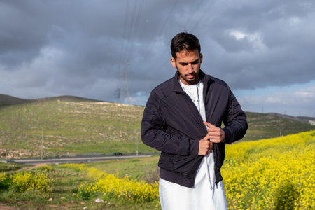 A man in a black jacket and dishdasha stands in a vibrant field of yellow flowers and green hills,...
