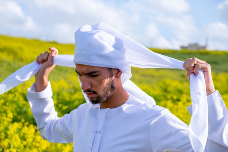 A man in a white dishdasha and kandura stands in a field of yellow flowers, lifting the kandora with eyes closed, enjoying the spring sunshine, surrounded by green hills under a partly cloudy skyの写真素材