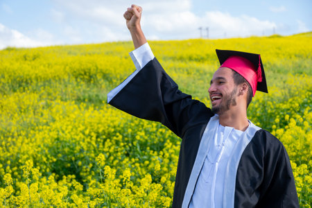 Cheerful young male wearing kandora and celebrating his graduation in a beautiful yellow flowers field with smile on his face rising his fist to the skyの写真素材