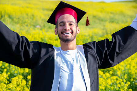 Cheerful young male wearing kandora and celebrating his graduation in a beautiful yellow flowers field with smile on his face rising his fist to the skyの写真素材