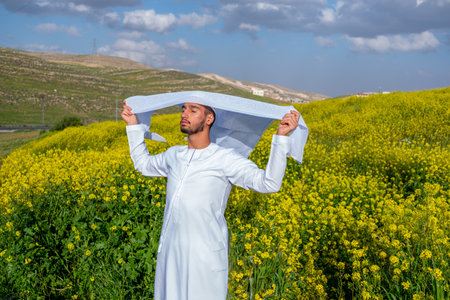 A man in a white dishdasha and kandura stands in a field of yellow flowers, lifting the kandora with eyes closed, enjoying the spring sunshine, surrounded by green hills under a partly cloudy skyの写真素材