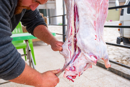 The butcher is cutting meat outdoors, holding his knife, and smiling with a bearded face