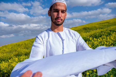 A man in a white dishdasha and kandura stands in a field of yellow flowers, lifting the kandora with...