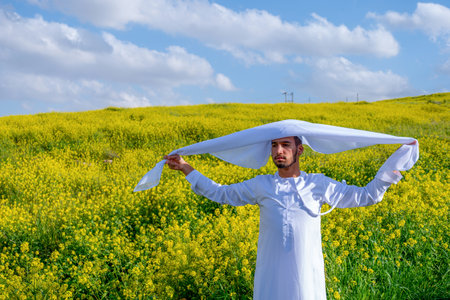 A man in a white dishdasha and kandura stands in a field of yellow flowers, lifting the kandora with eyes closed, enjoying the spring sunshine, surrounded by green hills under a partly cloudy skyの写真素材