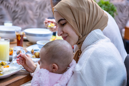 A tender portrait captures a mother seated at the family table, lovingly caring for her baby while they share a meal together. The bond between them shines through as they create cherished memories in the heart of their homeの写真素材