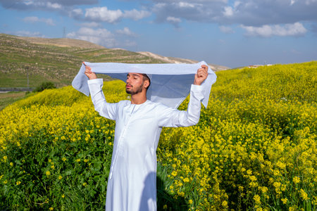 A man in a white dishdasha and kandura stands in a field of yellow flowers, lifting the kandora with eyes closed, enjoying the spring sunshine, surrounded by green hills under a partly cloudy skyの写真素材