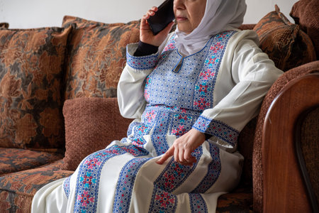Female wearing palestinian traditional clothes indoor making phone call on couch  with her relatives in palestineの写真素材