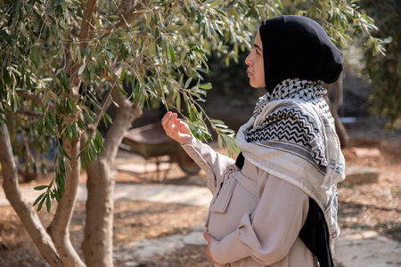 Female holding branch of olive tree while wearing palestinian keffiyeh in the fieldの写真素材
