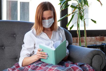 Woman in medical mask reading a book while sitting on a sofaの写真素材
